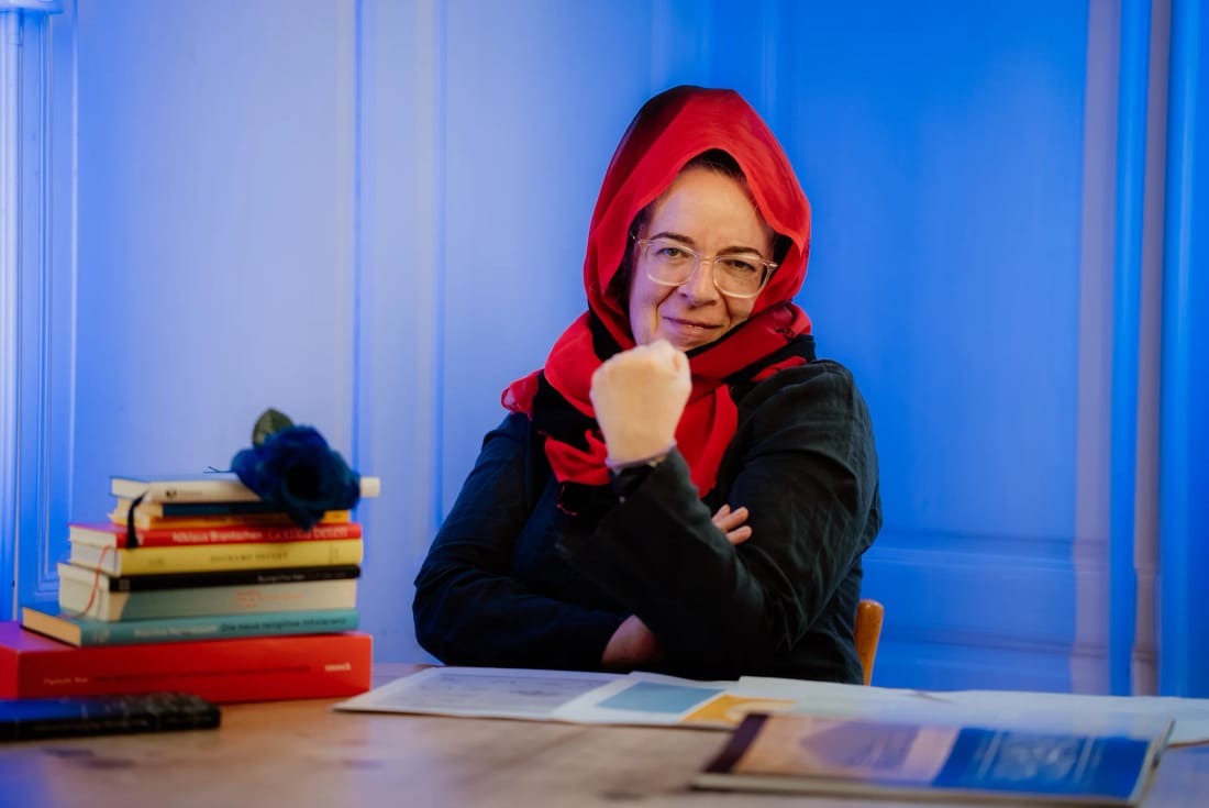 Pfarrerin am Set von «Hä? Gott!» vor blauer Wand mit Bücherstapel